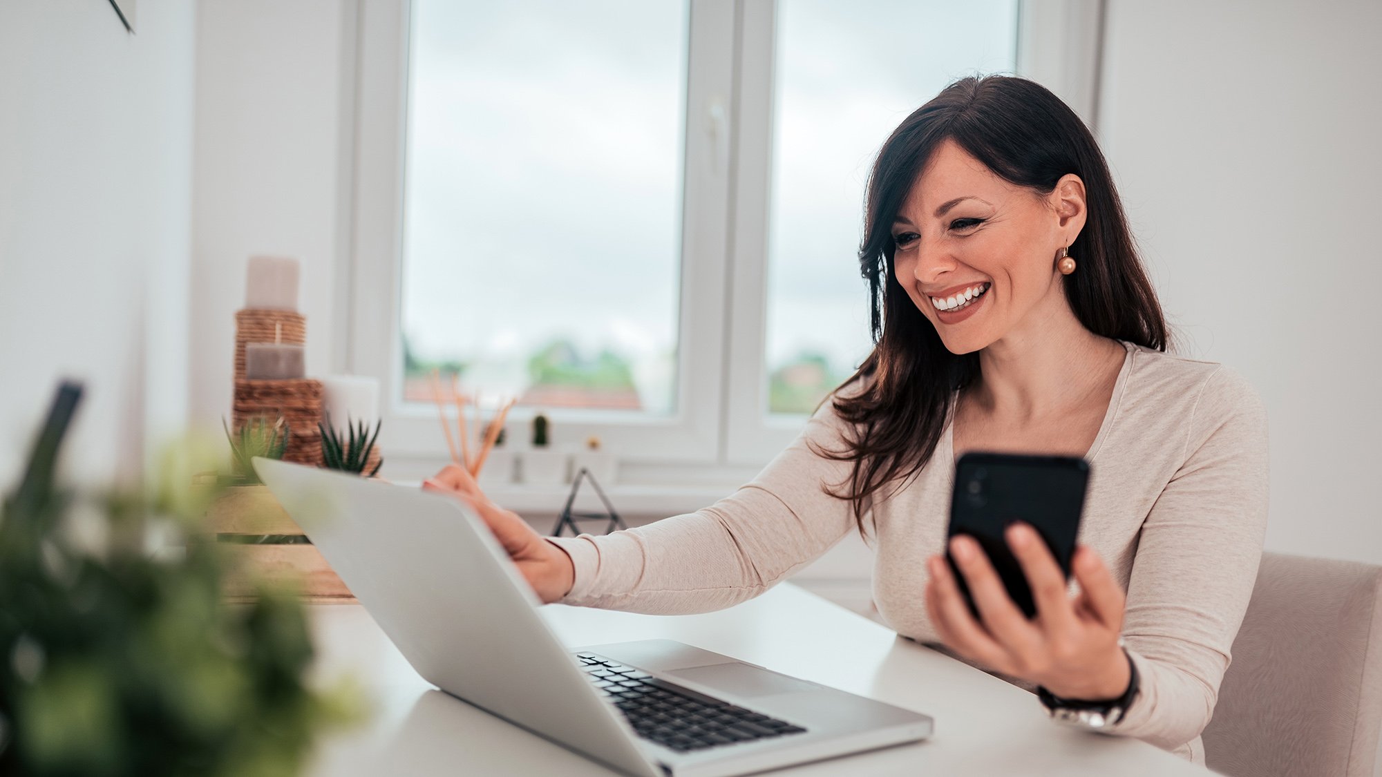 Een vrouw multitaskt met haar telefoon en laptop op haar thuiswerkplek